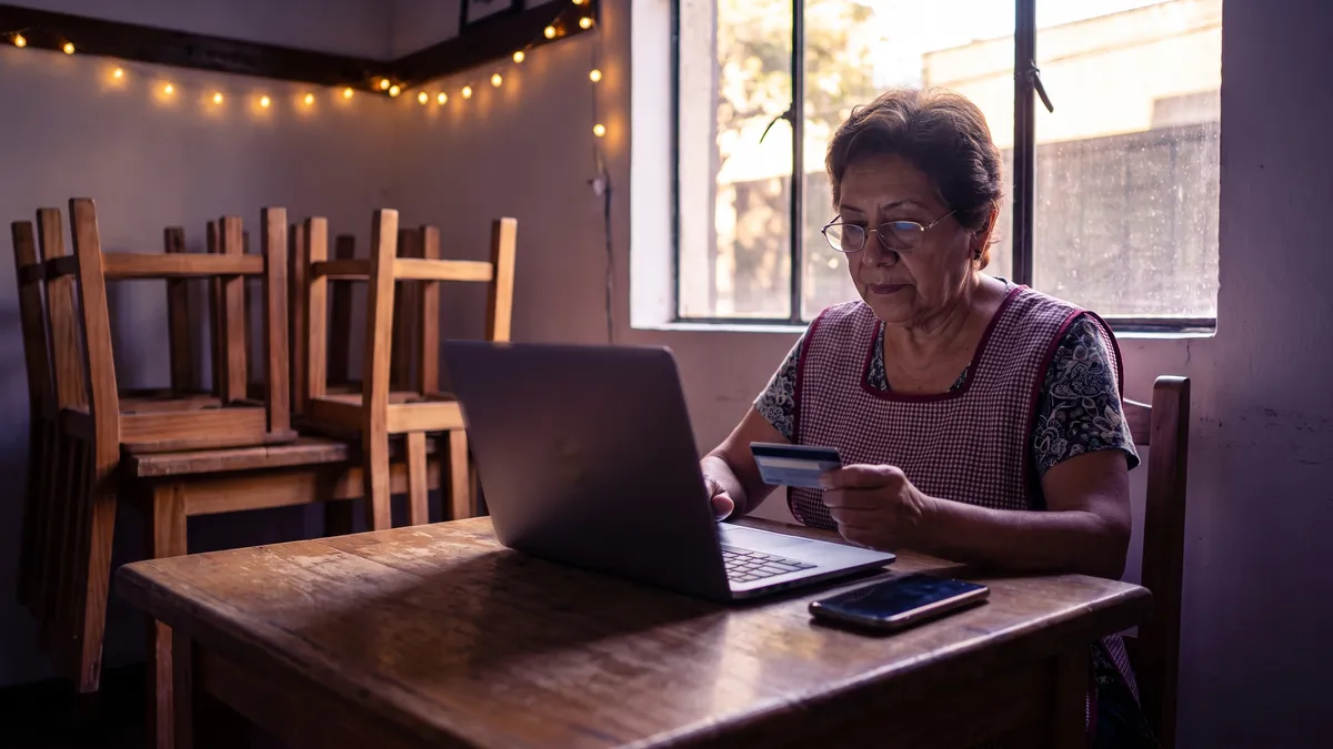 The owner of a coffee shop, already closed for the day, reviews payment settings on a laptop with a card in hand and his phone beside him